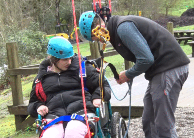 A young person in a wheel chair is being supported to put on a harness ready to do high rope climbing