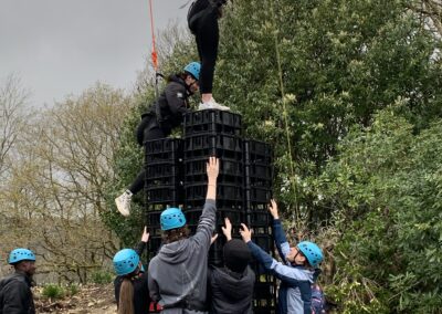 A group of delegates in safety helmets build a tower of crates using co operation and communication skills. The tower is very high, the young people on top of the tower are kept safe by high ropes.