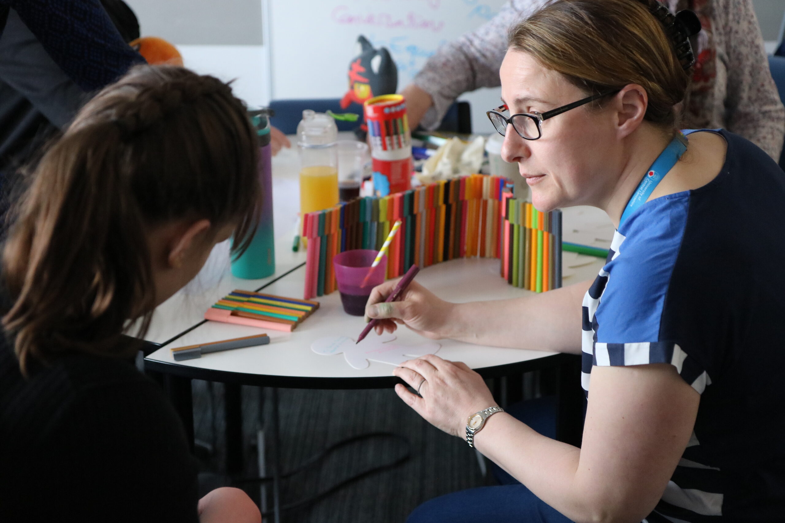 A You Can facilitator supports a young person to tell her ideas in the Don't Judge a Book by Its Cover young people led project. There are coloured pens on the table and paper.