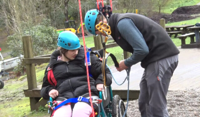 A young person in a wheel chair is being supported to put on a harness ready to do high rope climbing