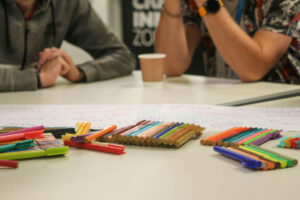 A young person and facilitator in UCan are discussing some ideas that have been written on a long piece of paper. There are lots of coloured pens in the foreground.