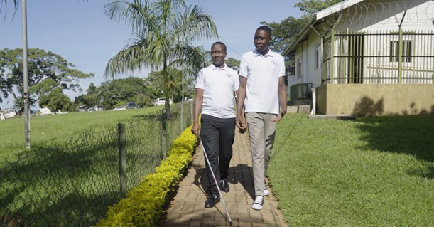 Two youth researchers in Uganda. One is visually impaired and carrying a white stick. The other is holding his hand. They are walking past a green grass area.
