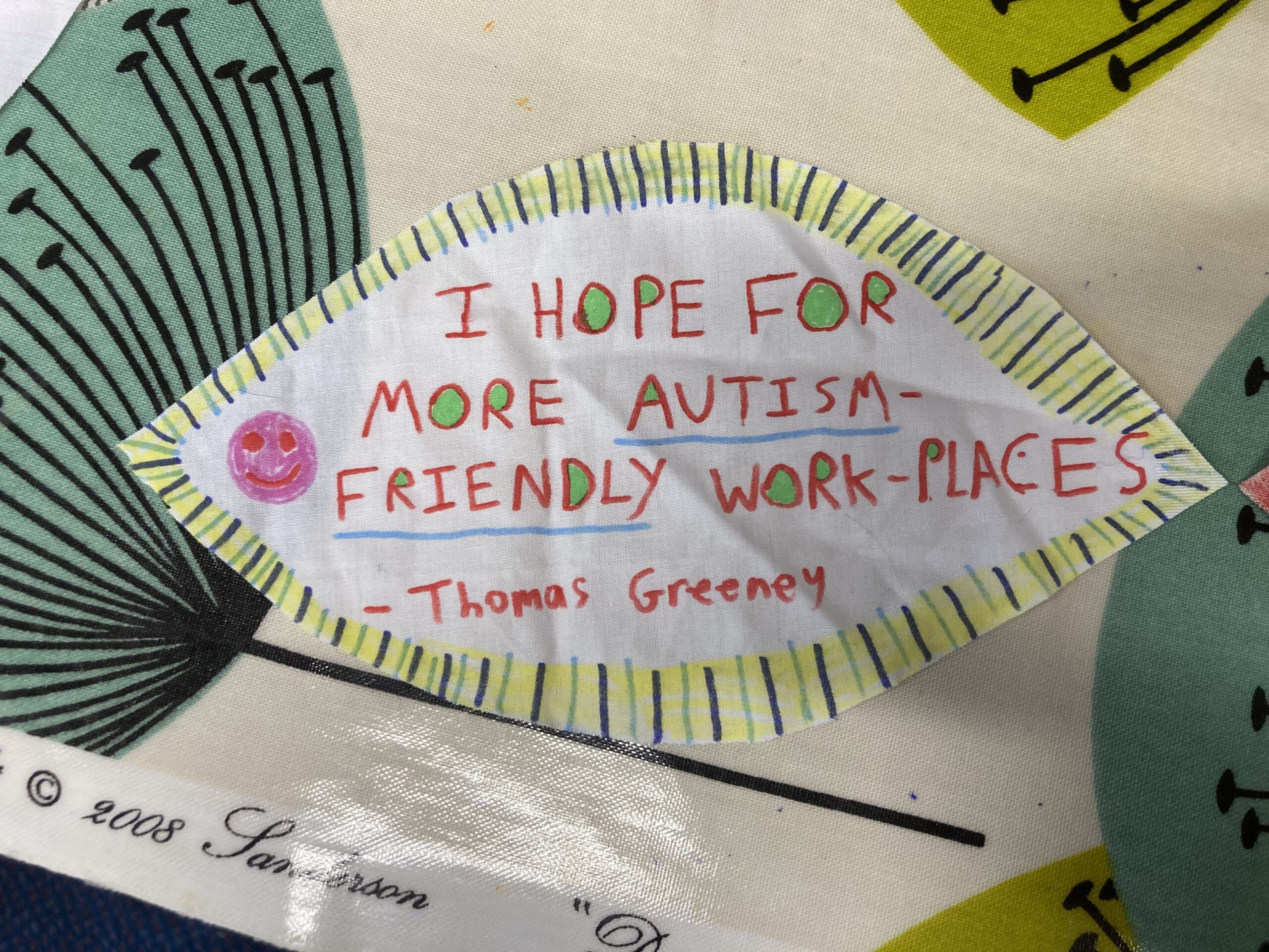 The image is of a piece of paper shaped like an eye. The writing in the middle reads "I hope for more autism friendly work-places". There is a pink smiley face in the corner and the border is yellow, green and blue.